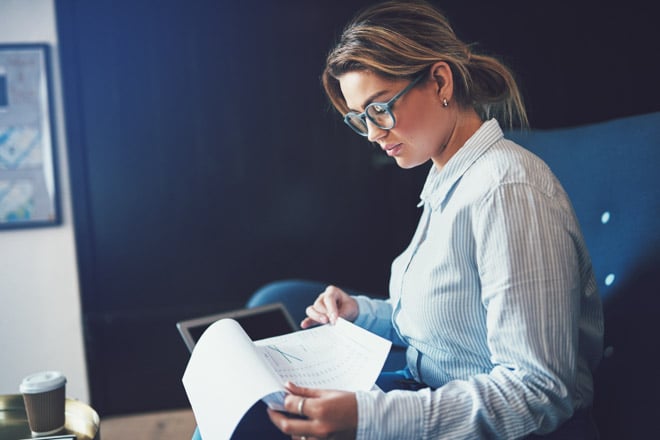 Young businesswoman sitting on a sofa at home reading through paperwork and using a laptop